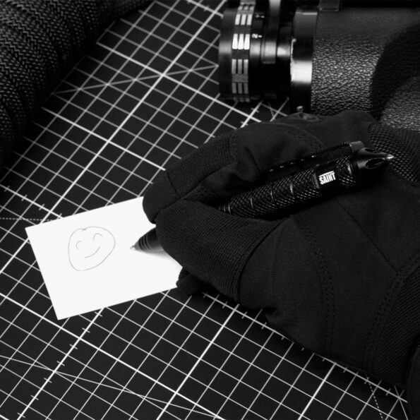 Gloved hand using the matte black SAINT tactical pen to write on a card